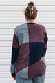 A woman viewed from behind wearing a color-block knit sweater with panels of burgundy, navy, and light blue-gray, paired with light-wash jeans against a white siding background.