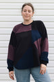 A woman wearing a color-block knit sweater with geometric panels of black, navy blue, and heathered purple, paired with light-wash blue jeans, standing against a white siding background.