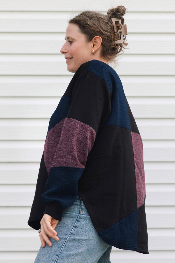 A woman wearing a color-block knit cardigan with navy, black, and heathered mauve panels, paired with light-wash blue jeans, standing against a white horizontal siding wall.