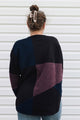 A woman viewed from behind wearing a color-block knit sweater with large panels of black, navy, and heathered plum purple, paired with blue jeans against a white siding background.