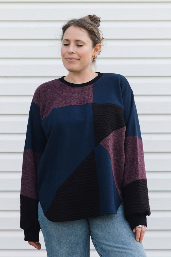 A woman wearing a color-block knit sweater with navy, burgundy, and black panels, paired with light blue jeans, standing against a white siding background.