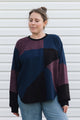 A woman wearing a color-block knit sweater with navy, burgundy, and black panels, paired with light blue jeans, standing against a white siding background.