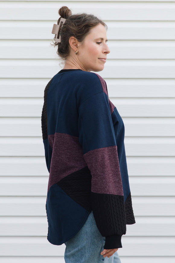A woman shown from the side profile wearing a color-block knit sweater with navy, burgundy, and black panels, paired with light blue jeans, standing against a white horizontal siding background.