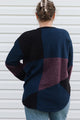 A woman viewed from behind wearing a patchwork knit sweater with navy, black, and burgundy sections, paired with blue jeans in front of a white siding background.