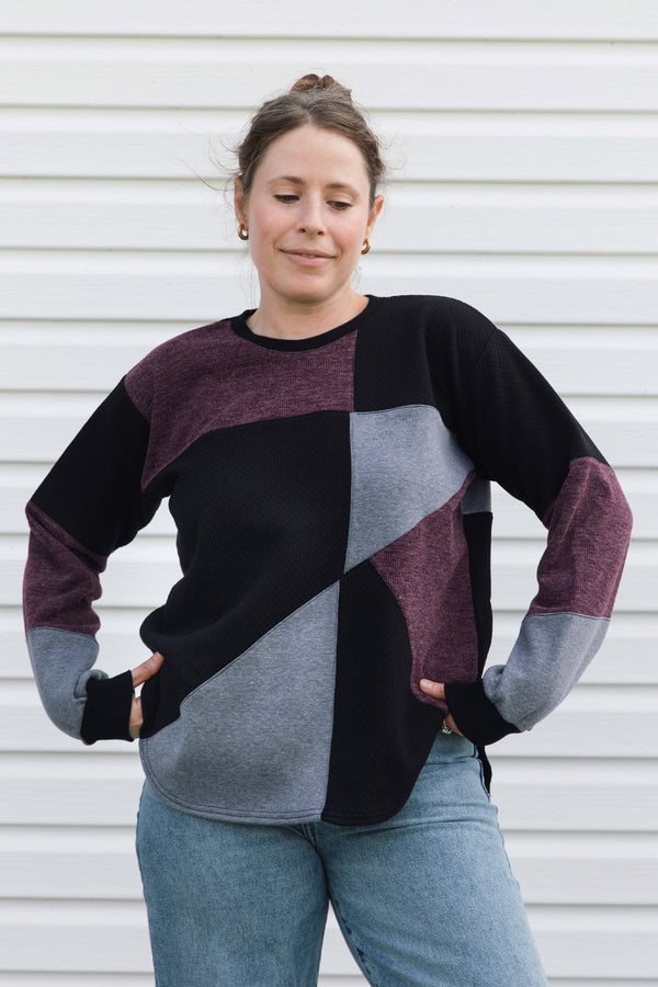 A woman wearing a color-block knit sweater with black, heather gray, and heathered plum sections, paired with blue jeans, standing against a white siding background.