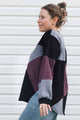 A woman wearing a color-block knit sweater with black, gray, and burgundy panels, paired with blue jeans, standing against a white horizontal siding background.