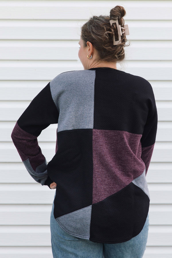 A woman viewed from behind wearing a color-block knit sweater with panels of black, heather gray, and heathered plum purple, paired with blue jeans against a white siding background.