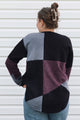 A woman viewed from behind wearing a color-block knit sweater with panels of black, heather gray, and heathered plum purple, paired with blue jeans against a white siding background.