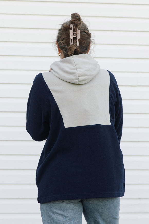 A person viewed from behind wearing a color-block hoodie with navy blue and light gray panels, paired with light blue jeans, standing against a white siding wall.