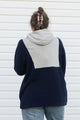 A person viewed from behind wearing a color-block hoodie with navy blue and light gray panels, paired with light blue jeans, standing against a white siding wall.