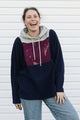 A woman laughing while wearing a color-blocked hoodie with a navy blue body, a gray hood, and a maroon chest panel, paired with light blue jeans, standing against a white siding background.