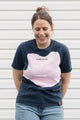 Woman wearing a navy blue and pink t-shirt with text, standing against a white wall.