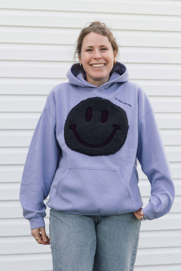 A woman wearing a lavender hoodie with a large black fuzzy smiley face patch on the front, standing against a white siding wall.