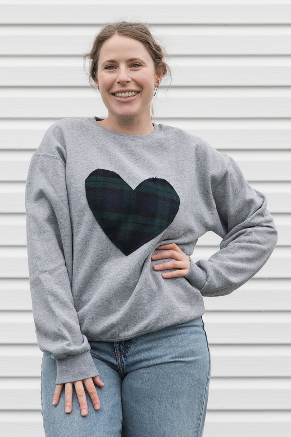 Woman wearing a unisex light grey sweatshirt with a navy, green plaid heart. 