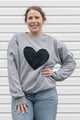 Woman wearing a unisex light grey sweatshirt with a navy, green plaid heart. 