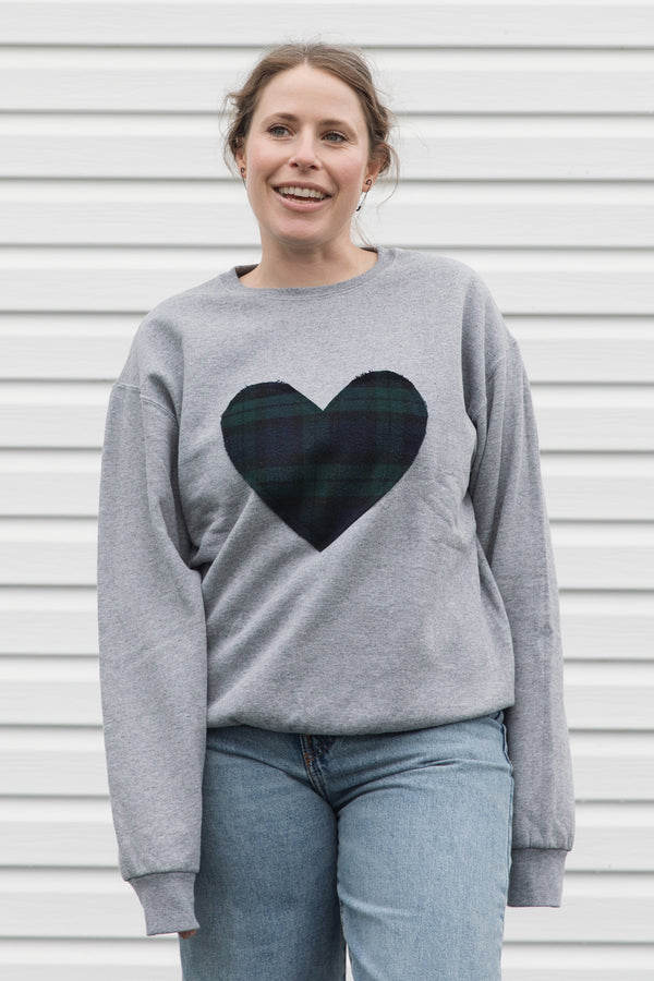 Woman wearing a unisex light grey sweatshirt with a navy, green plaid heart. 