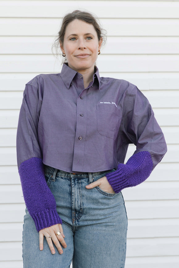 A woman wearing a purple button-down shirt with purple knit elbow-length sleeves and blue jeans, standing against a white siding background.
