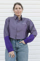 A woman wearing a purple button-down shirt with purple knit elbow-length sleeves and blue jeans, standing against a white siding background.