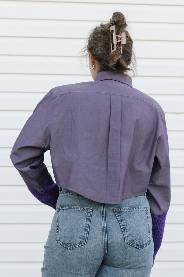 A woman viewed from behind wearing a purple long-sleeve button-up shirt with a unique back seam and purple knit cuffs, paired with blue jeans, set against a white paneled wall.