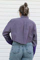 A woman viewed from behind wearing a purple long-sleeve button-up shirt with a unique back seam and purple knit cuffs, paired with blue jeans, set against a white paneled wall.