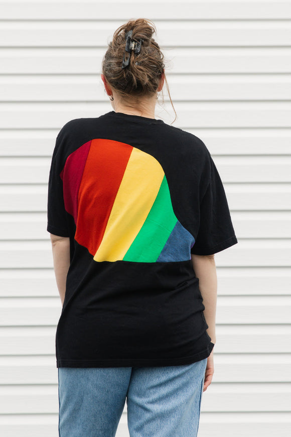 Person wearing a black t-shirt with a rainbow design on the back against a white wall.