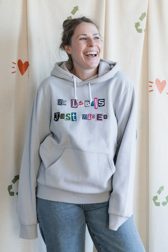 Person wearing a gray hoodie with colorful text, reading 'no labels, just vibes.' upcycled in canada