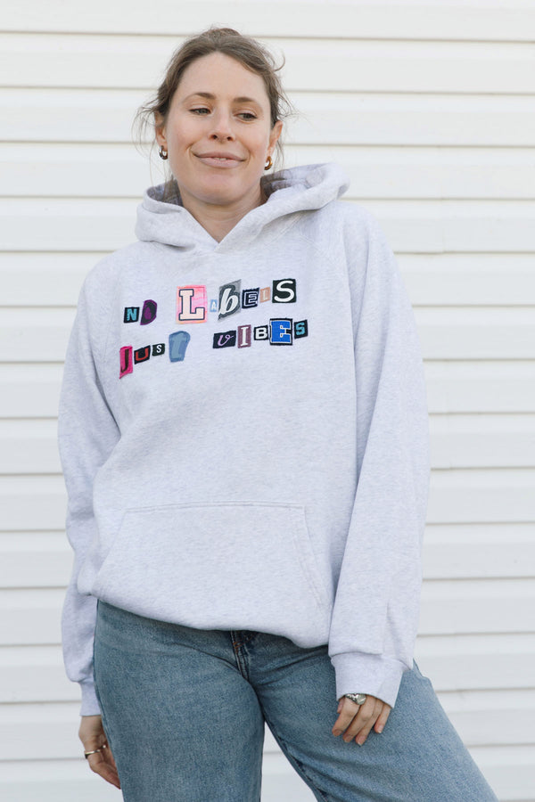 A woman wearing a light heather gray hoodie with colorful embroidered letters spelling 'LOVE JOY' on the front, paired with blue jeans, standing against a white paneled wall.