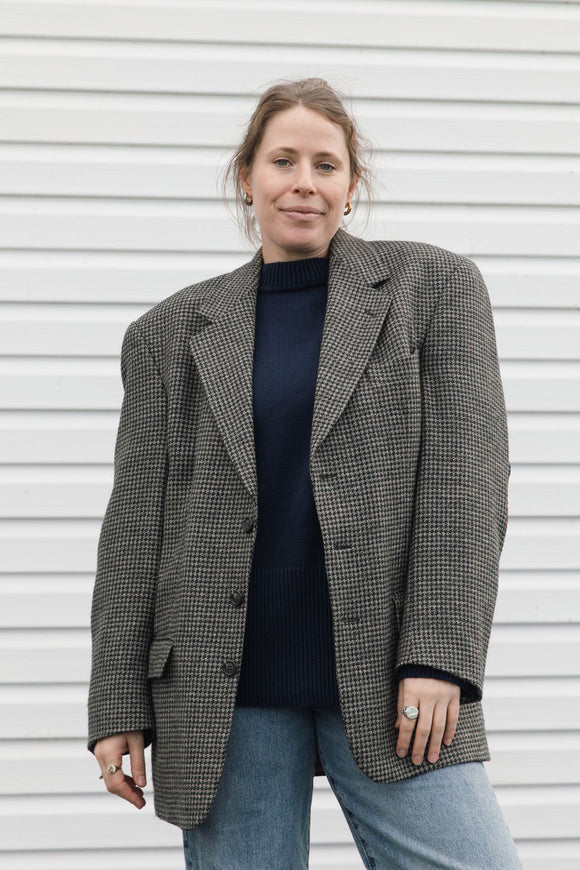 A woman wearing an oversized black and white houndstooth blazer over a dark navy turtleneck sweater and light blue jeans, standing against a white horizontal-slat wall.