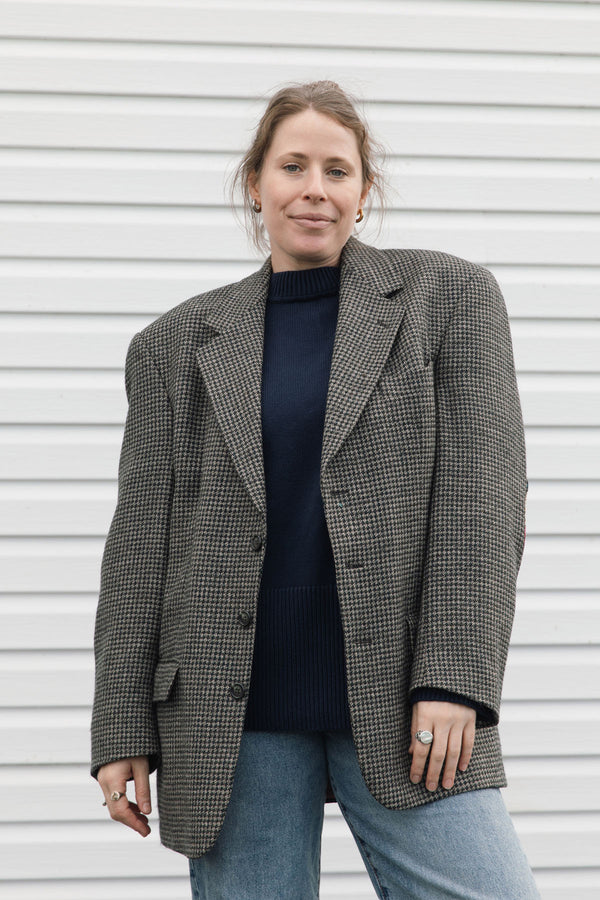 A woman wearing an oversized black and white houndstooth blazer over a dark navy turtleneck sweater and light blue jeans, standing against a white horizontal-slat wall.