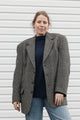 A woman wearing an oversized black and white houndstooth blazer over a dark navy turtleneck sweater and light blue jeans, standing against a white horizontal-slat wall.