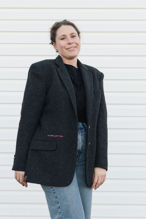 Person wearing a dark gray blazer with text on it, standing against a white wall.