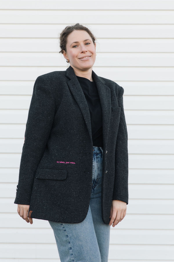 Person wearing a dark gray blazer with text on it, standing against a white wall.