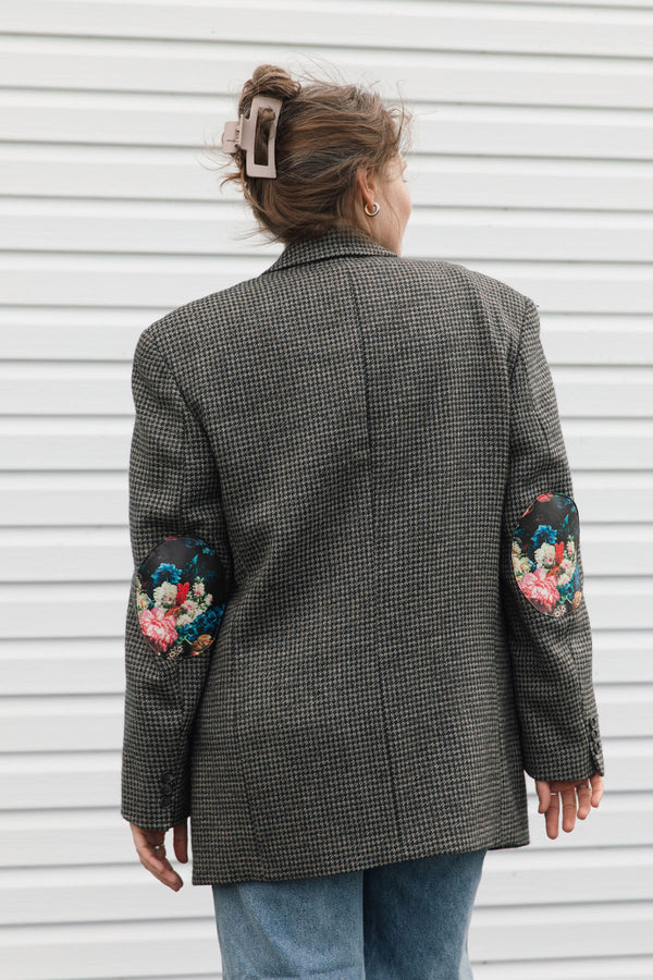 A woman viewed from behind wearing a dark gray and black houndstooth blazer with colorful floral embroidery on the sleeves, paired with blue jeans, standing against a white siding background.