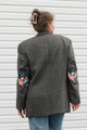 A woman viewed from behind wearing a dark gray and black houndstooth blazer with colorful floral embroidery on the sleeves, paired with blue jeans, standing against a white siding background.