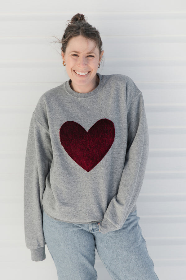 Person wearing a gray sweater with a red velvet heart design against a white background, sewn in canada