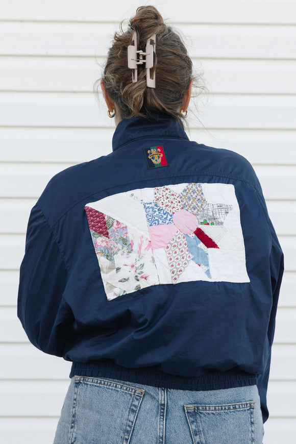 A person viewed from behind wearing a navy blue bomber jacket with a large multicolor patchwork quilt design on the back, paired with light blue jeans against a white siding background.