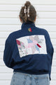 A person viewed from behind wearing a navy blue bomber jacket with a large multicolor patchwork quilt design on the back, paired with light blue jeans against a white siding background.